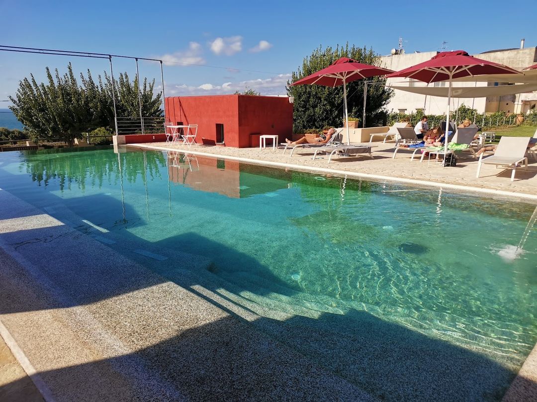 The hotel's sparkling turquoise pool with red umbrellas, loungers, and a red building structure overlooking Mediterranean views.