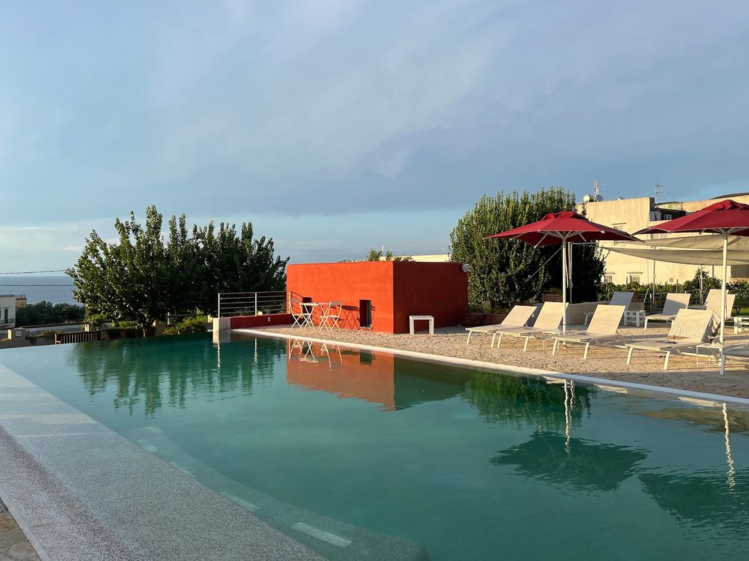 The hotel's turquoise pool with loungers, red umbrellas, and a modern orange structure reflected in the water at sunset.